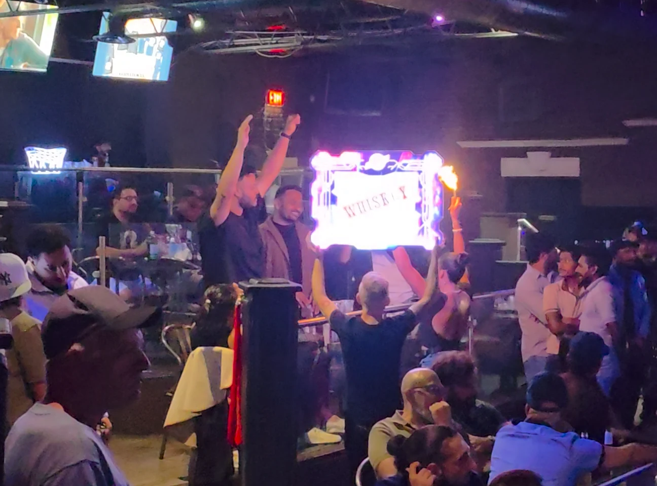 Guests raise their hands as Whiskey bottle service arrives with sparklers and an illuminated sign.