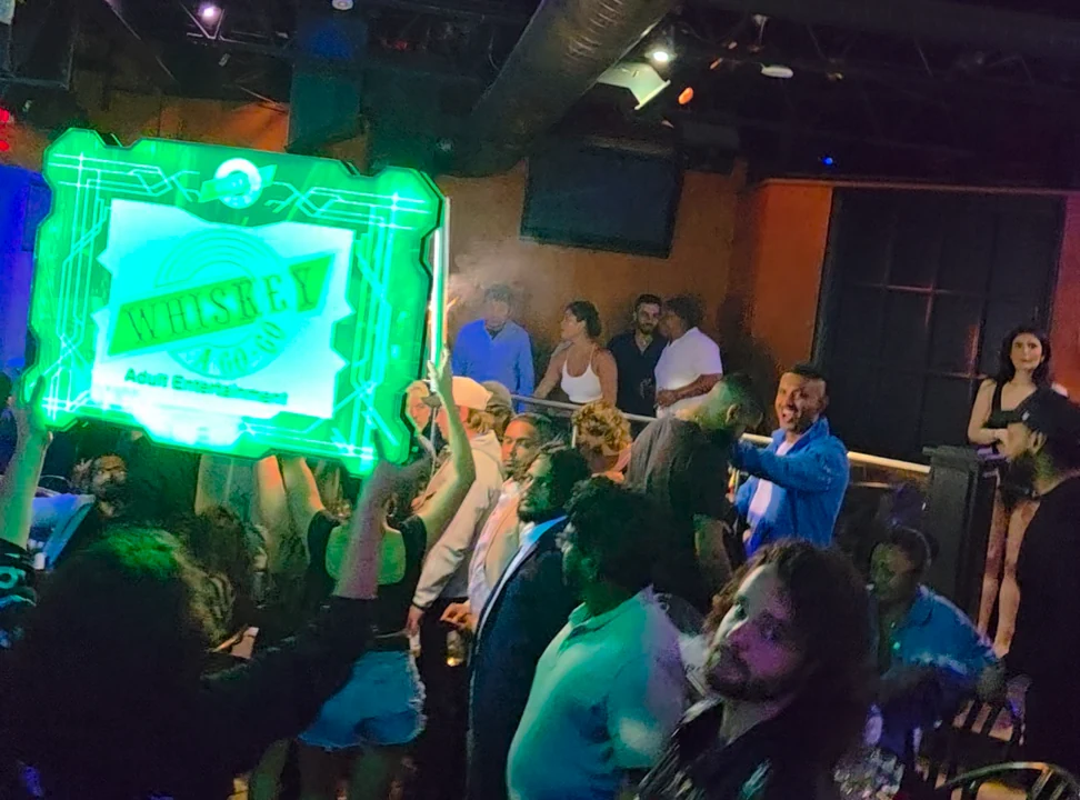 Guests smile and cheer beside an illuminated Whiskey sign as bottle service reaches their booth.